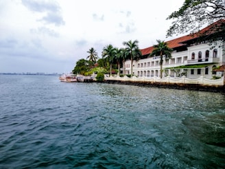 A coastal scene featuring a large, white colonial-style building with a red roof, framed by several tall palm trees and lush greenery. The structure stands adjacent to a calm body of water, with a small dock extending into the water where a couple of boats are moored. The sky appears overcast, creating a tranquil and relaxed atmosphere.