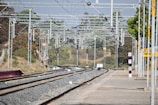A railway station's tracks extend into the distance under a network of overhead electrical wires. Signal lights and signs are visible along the tracks, with trees and vegetation alongside. A platform runs parallel to the tracks, with a sign reading 'VTO' on it.
