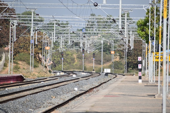 A railway station's tracks extend into the distance under a network of overhead electrical wires. Signal lights and signs are visible along the tracks, with trees and vegetation alongside. A platform runs parallel to the tracks, with a sign reading 'VTO' on it.