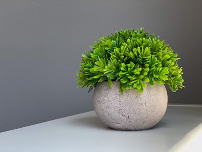 A round, textured pot holds a lush, green artificial plant on a white surface. The background is a smooth gray wall, and the lighting casts gentle shadows on the surface.