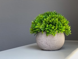 A round, textured pot holds a lush, green artificial plant on a white surface. The background is a smooth gray wall, and the lighting casts gentle shadows on the surface.