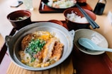 A hearty meal is served in a metal pan, featuring a fried cutlet covered in sauce, surrounded by scrambled eggs and topped with chopped green onions. Adjacent to the dish are bowls filled with rice and a green soup, placed on red lacquered trays. Chopsticks rest on the edge of the rice bowl, while a light blue spoon sits on an empty dish.