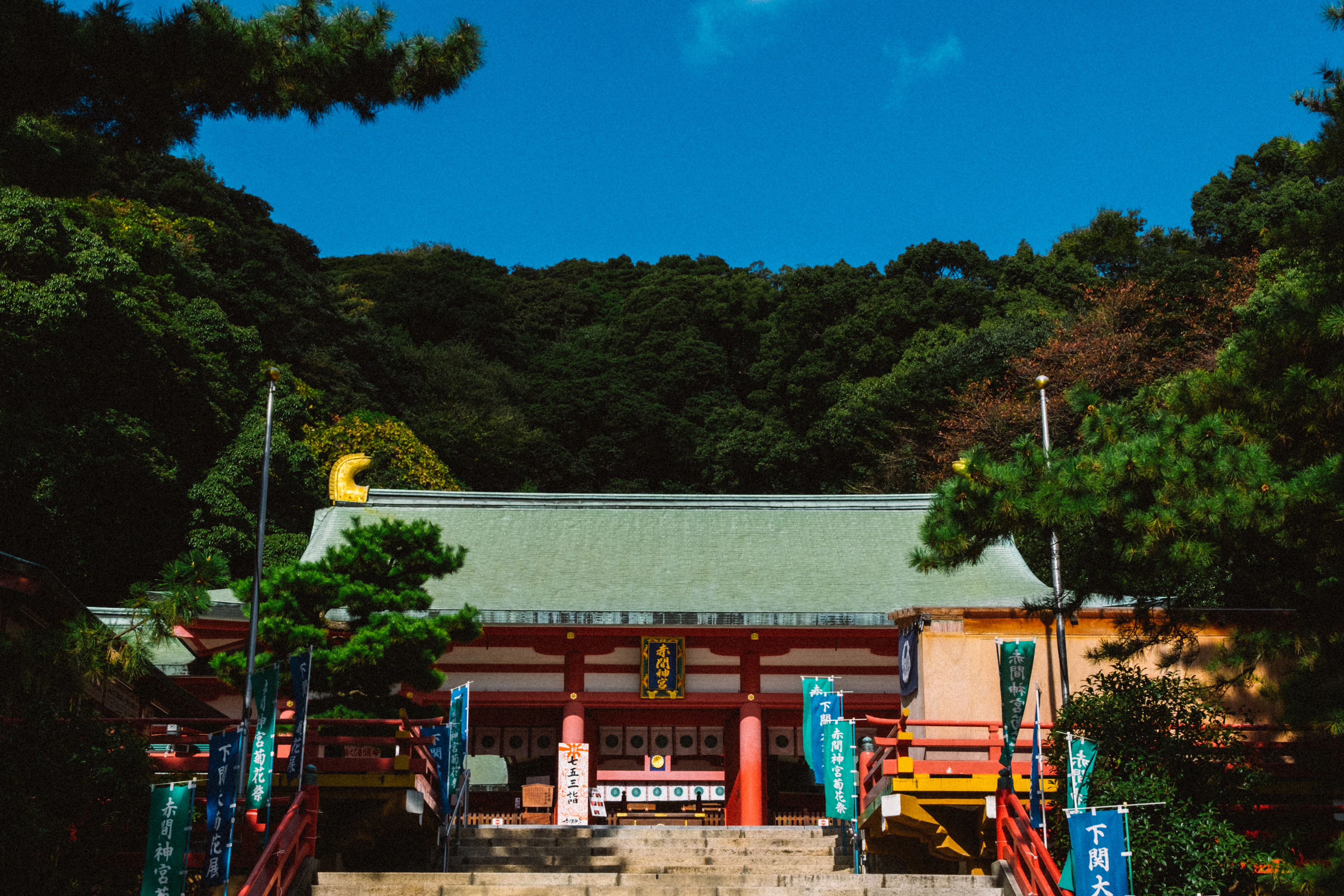 A red-and-white shrine hall hung with banners stands atop stone steps under a clear blue sky and green trees.