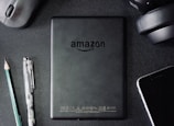 A flat lay composition on a dark textured surface includes a black Amazon Kindle device, surrounded by a pencil, a pen, a computer mouse, a pair of headphones, and the corner of a smartphone. The Kindle's back is centered and showcases the Amazon logo.