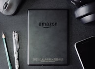 A flat lay composition on a dark textured surface includes a black Amazon Kindle device, surrounded by a pencil, a pen, a computer mouse, a pair of headphones, and the corner of a smartphone. The Kindle's back is centered and showcases the Amazon logo.