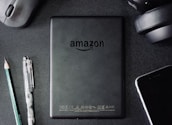 A flat lay composition on a dark textured surface includes a black Amazon Kindle device, surrounded by a pencil, a pen, a computer mouse, a pair of headphones, and the corner of a smartphone. The Kindle's back is centered and showcases the Amazon logo.