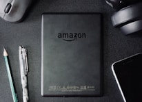 A flat lay composition on a dark textured surface includes a black Amazon Kindle device, surrounded by a pencil, a pen, a computer mouse, a pair of headphones, and the corner of a smartphone. The Kindle's back is centered and showcases the Amazon logo.