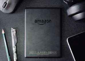 A flat lay composition on a dark textured surface includes a black Amazon Kindle device, surrounded by a pencil, a pen, a computer mouse, a pair of headphones, and the corner of a smartphone. The Kindle's back is centered and showcases the Amazon logo.