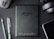 A flat lay composition on a dark textured surface includes a black Amazon Kindle device, surrounded by a pencil, a pen, a computer mouse, a pair of headphones, and the corner of a smartphone. The Kindle's back is centered and showcases the Amazon logo.