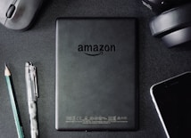 A flat lay composition on a dark textured surface includes a black Amazon Kindle device, surrounded by a pencil, a pen, a computer mouse, a pair of headphones, and the corner of a smartphone. The Kindle's back is centered and showcases the Amazon logo.