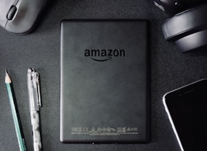 A flat lay composition on a dark textured surface includes a black Amazon Kindle device, surrounded by a pencil, a pen, a computer mouse, a pair of headphones, and the corner of a smartphone. The Kindle's back is centered and showcases the Amazon logo.