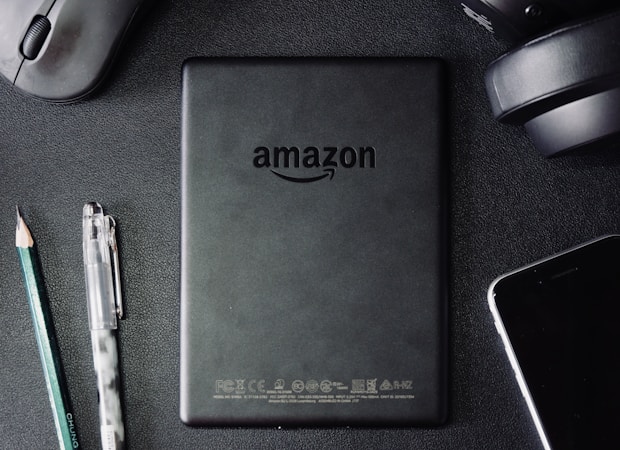 A flat lay composition on a dark textured surface includes a black Amazon Kindle device, surrounded by a pencil, a pen, a computer mouse, a pair of headphones, and the corner of a smartphone. The Kindle's back is centered and showcases the Amazon logo.