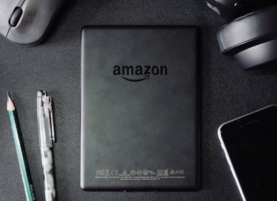 A flat lay composition on a dark textured surface includes a black Amazon Kindle device, surrounded by a pencil, a pen, a computer mouse, a pair of headphones, and the corner of a smartphone. The Kindle's back is centered and showcases the Amazon logo.