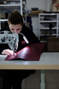 A person is focused on stitching a piece of red fabric using a sewing machine in a workshop. The setting appears organized with shelves in the background holding various items. The person is wearing a dark hoodie, and the workspace is well-lit with a lamp attached to the machine.