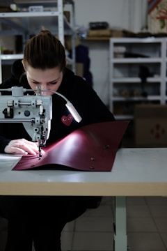 A person is focused on stitching a piece of red fabric using a sewing machine in a workshop. The setting appears organized with shelves in the background holding various items. The person is wearing a dark hoodie, and the workspace is well-lit with a lamp attached to the machine.