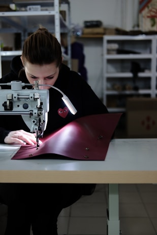 A person is focused on stitching a piece of red fabric using a sewing machine in a workshop. The setting appears organized with shelves in the background holding various items. The person is wearing a dark hoodie, and the workspace is well-lit with a lamp attached to the machine.
