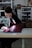 A person is focused on stitching a piece of red fabric using a sewing machine in a workshop. The setting appears organized with shelves in the background holding various items. The person is wearing a dark hoodie, and the workspace is well-lit with a lamp attached to the machine.