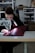 A person is focused on stitching a piece of red fabric using a sewing machine in a workshop. The setting appears organized with shelves in the background holding various items. The person is wearing a dark hoodie, and the workspace is well-lit with a lamp attached to the machine.