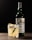 A glass with a whiskey cocktail, garnished with a sprig of rosemary and lemon peel, sits on a wooden surface. Behind it, there is a bottle of whiskey with a green tint and a white label.