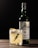 A glass with a whiskey cocktail, garnished with a sprig of rosemary and lemon peel, sits on a wooden surface. Behind it, there is a bottle of whiskey with a green tint and a white label.