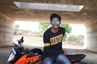 A person wearing sunglasses and a black sweater with 'REBEL' written on it sits confidently on an orange motorcycle under a concrete structure. There is greenery and rocky terrain visible in the background through the opening.