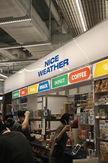 A busy indoor stall with various food sections including grocery, deli, bakery, snack, donut, and coffee. People are interacting at the counter, some holding beverages and wearing masks.