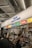 A busy indoor stall with various food sections including grocery, deli, bakery, snack, donut, and coffee. People are interacting at the counter, some holding beverages and wearing masks.
