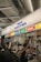 A busy indoor stall with various food sections including grocery, deli, bakery, snack, donut, and coffee. People are interacting at the counter, some holding beverages and wearing masks.