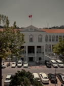A historical building with a symmetrical facade, featuring several large windows and a prominent entrance with columns. A red flag with a crescent and star is prominently displayed on the rooftop. Several cars are parked in front, with people gathered around. The surrounding environment includes large trees and a minaret visible in the distance.