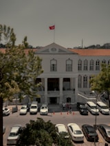 A historical building with a symmetrical facade, featuring several large windows and a prominent entrance with columns. A red flag with a crescent and star is prominently displayed on the rooftop. Several cars are parked in front, with people gathered around. The surrounding environment includes large trees and a minaret visible in the distance.