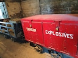 A wooden box painted red with the words 'DANGER EXPLOSIVES' printed in white. It is secured with a padlock and placed on a metal cart with four wheels. The cart is on a concrete floor against a rough brick wall, suggesting an industrial or storage setting.