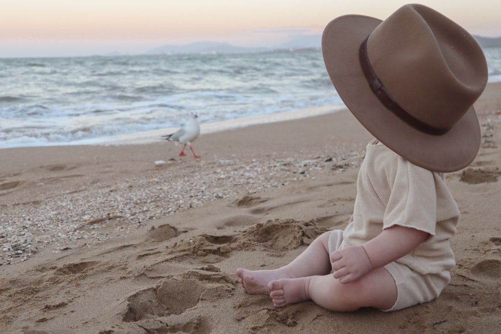child in white long sleeve shirt and brown hat sitting on brown sand beach during daytime