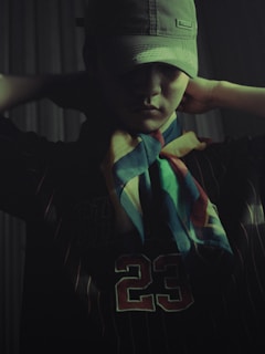 A person is wearing a cap that partially obscures their face, creating a shadowed effect. They are dressed in a dark-colored shirt with the number 23 visible, and have a multicolored scarf tied around their neck. Their arms are raised, with hands adjusting the cap.