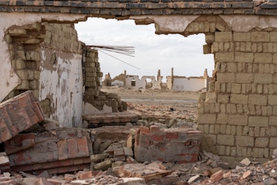 A scene of destruction with a partially collapsed building made of bricks and mud blocks. The foreground features broken bricks and debris, while through a large hole in the wall, more ruined buildings are visible in the background.
