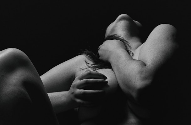 A black and white artistic photograph of a person with bare skin, featuring a dramatic play of shadows and light. The figure's pose is expressive and conveys a sense of introspection or emotion.