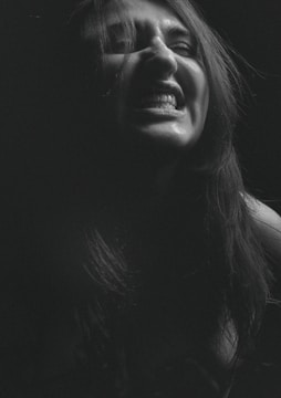 A person with long hair is captured in a dimly lit, dramatic black and white photograph, expressing an intense emotion with a wide-open mouth and visible teeth. The hair is partially covering the face, adding to the raw expression.