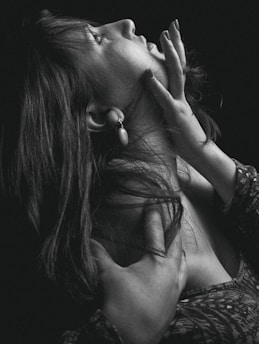A high-contrast black and white photograph of a woman in a dramatic pose, looking upwards. Her hair cascades over her shoulders, and her hands are elegantly placed around her face and neck, conveying a sense of introspection or contemplation.