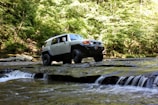 A rugged SUV is positioned on rocky terrain near a shallow, flowing river surrounded by dense green foliage. The vehicle is well-suited for off-road adventures, highlighting its durability and capability.