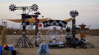 An artistic installation on a sandy beach featuring industrial-looking structures composed of metallic components, spherical elements, and a large circular element resembling a Ferris wheel. Some of the structures hold signs with text, and there are various mechanical and electronic parts throughout.