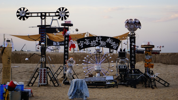 An artistic installation on a sandy beach featuring industrial-looking structures composed of metallic components, spherical elements, and a large circular element resembling a Ferris wheel. Some of the structures hold signs with text, and there are various mechanical and electronic parts throughout.