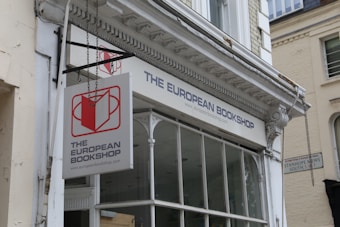The exterior of a bookshop with a prominent hanging sign displaying a red and white logo. The architecture features decorative molding and large windows. Nearby buildings have a classic, urban feel.