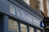 A storefront sign with the words 'la librairie' followed by 'Books in French Sold and Bought' in smaller text. The sign is mounted on a blue facade against a background of a brick building. A logo consisting of colorful vertical lines is also present on the sign.