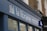 A storefront sign with the words 'la librairie' followed by 'Books in French Sold and Bought' in smaller text. The sign is mounted on a blue facade against a background of a brick building. A logo consisting of colorful vertical lines is also present on the sign.