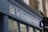 A storefront sign with the words 'la librairie' followed by 'Books in French Sold and Bought' in smaller text. The sign is mounted on a blue facade against a background of a brick building. A logo consisting of colorful vertical lines is also present on the sign.