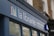 A storefront sign with the words 'la librairie' followed by 'Books in French Sold and Bought' in smaller text. The sign is mounted on a blue facade against a background of a brick building. A logo consisting of colorful vertical lines is also present on the sign.