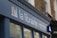 A storefront sign with the words 'la librairie' followed by 'Books in French Sold and Bought' in smaller text. The sign is mounted on a blue facade against a background of a brick building. A logo consisting of colorful vertical lines is also present on the sign.