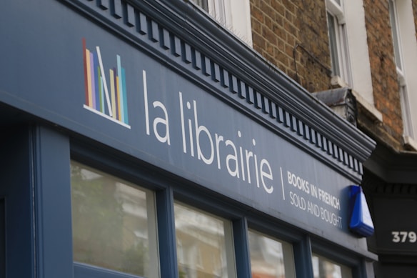 A storefront sign with the words 'la librairie' followed by 'Books in French Sold and Bought' in smaller text. The sign is mounted on a blue facade against a background of a brick building. A logo consisting of colorful vertical lines is also present on the sign.