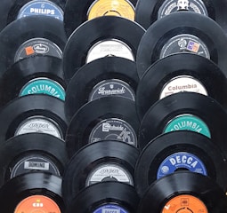 A collection of vintage vinyl records displayed in a pattern. Each record shows a different label from various recording companies such as Columbia, Decca, and Philips. The records have a glossy black finish and the labels feature a mix of colors including blue, orange, and white.