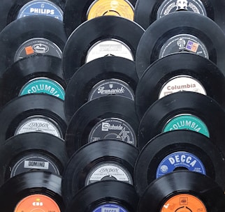 A collection of vintage vinyl records displayed in a pattern. Each record shows a different label from various recording companies such as Columbia, Decca, and Philips. The records have a glossy black finish and the labels feature a mix of colors including blue, orange, and white.