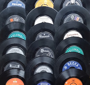 A collection of vintage vinyl records displayed in a pattern. Each record shows a different label from various recording companies such as Columbia, Decca, and Philips. The records have a glossy black finish and the labels feature a mix of colors including blue, orange, and white.